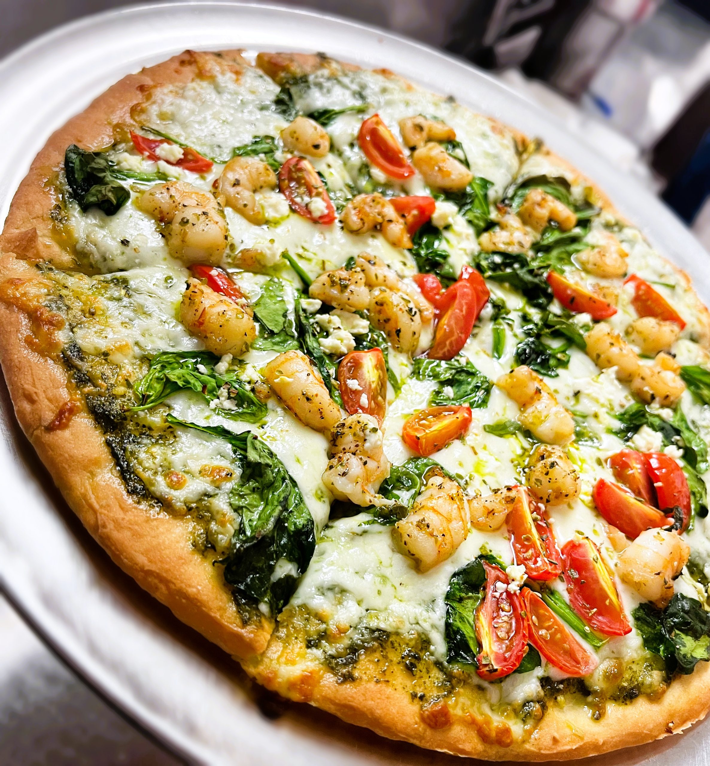 A pizza topped with shrimp, cherry tomatoes, spinach, and mozzarella cheese on a pesto sauce base.