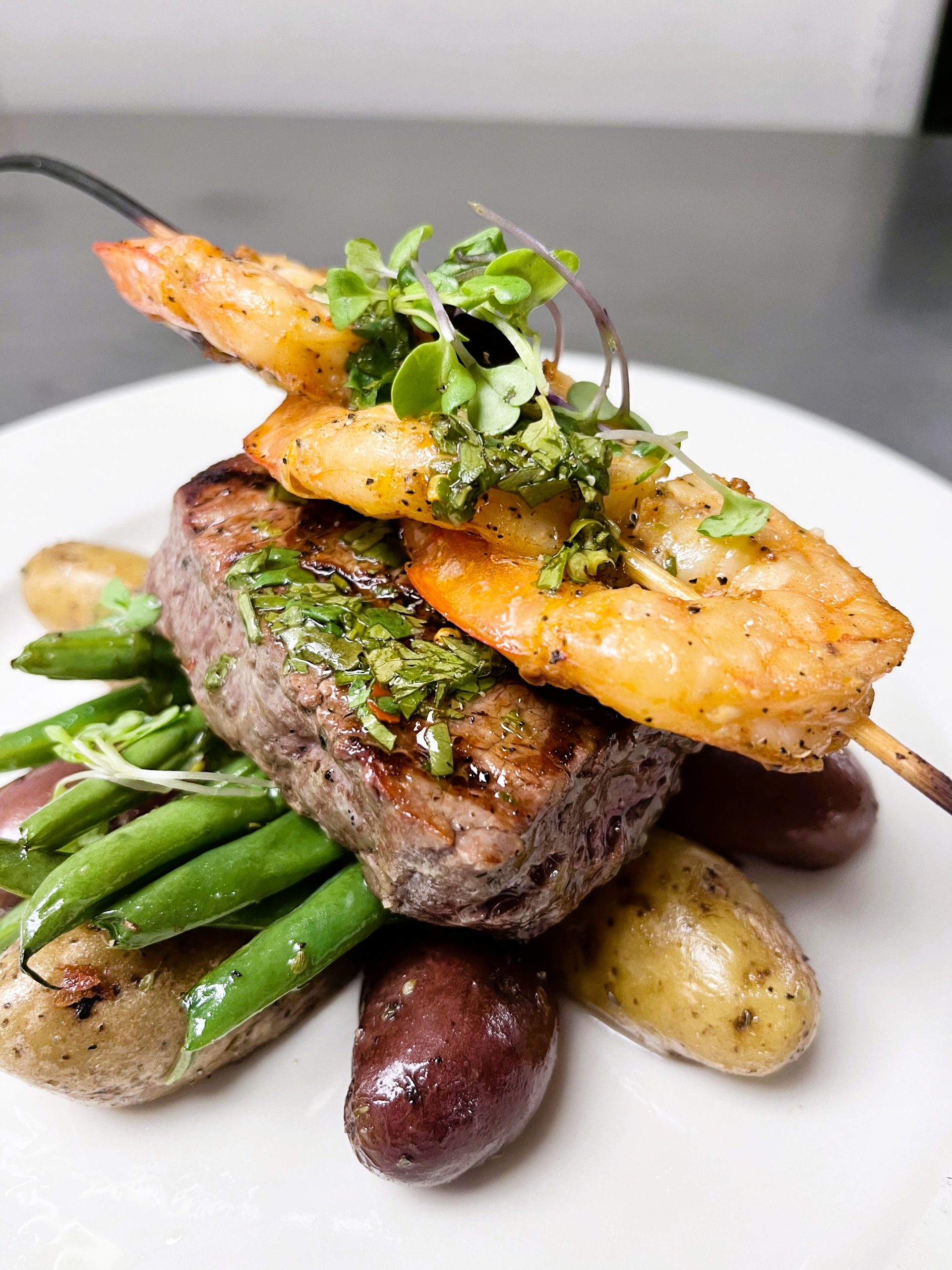 Steak topped with shrimp skewers, garnished with herbs, served on a bed of green beans and roasted potatoes.