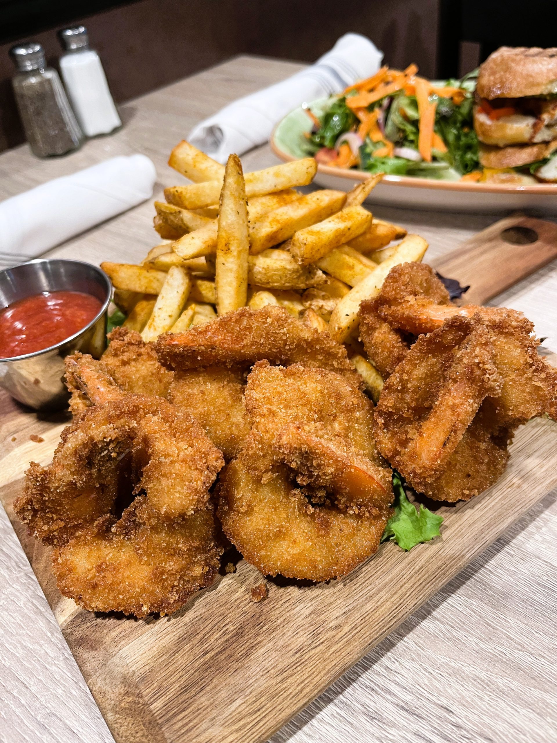 A plate of fried shrimp and fries with dipping sauce sits on a wooden board. A salad and sandwich are in the background.