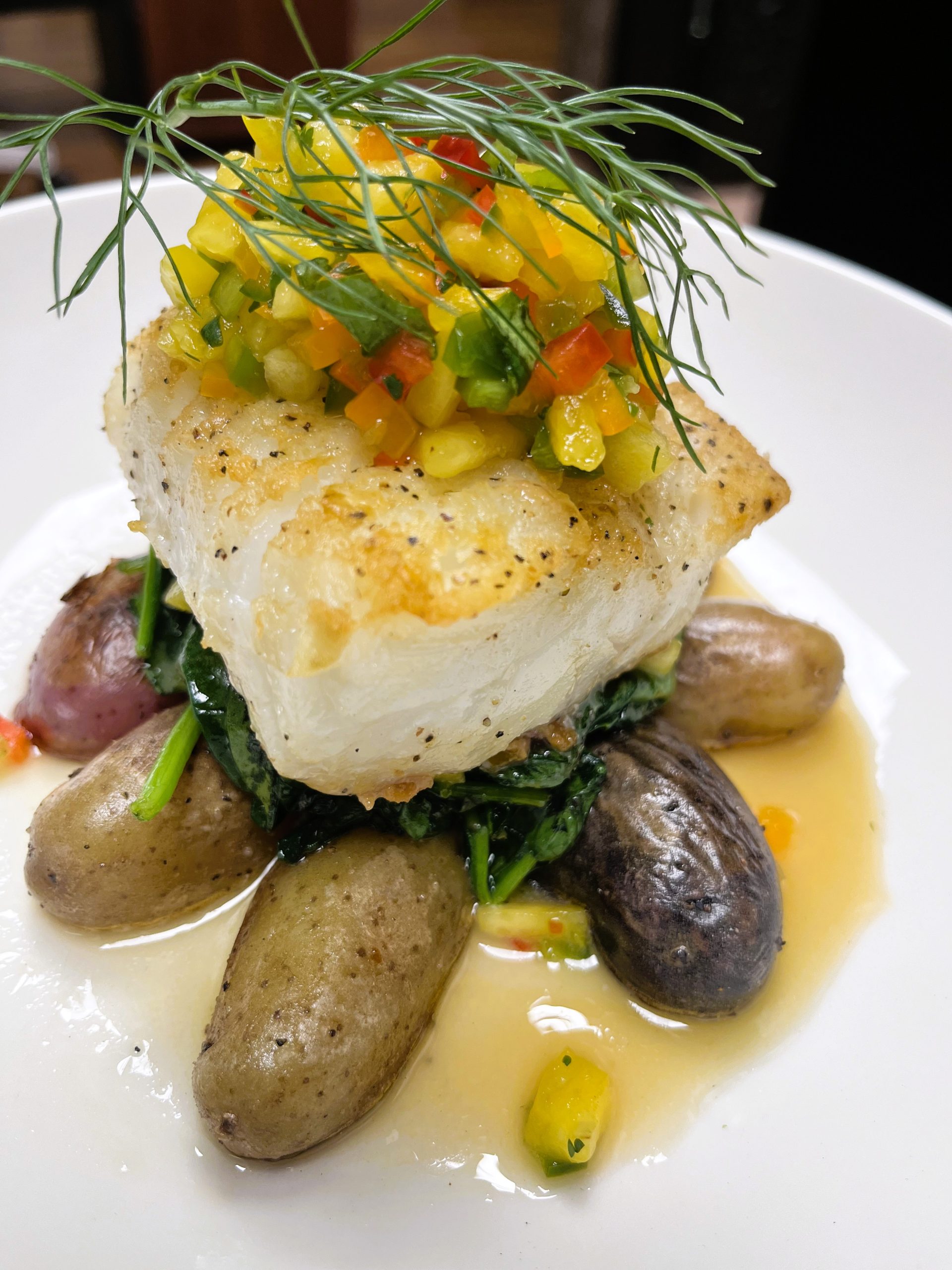 A seared fish fillet topped with diced bell peppers and herbs, served over spinach and fingerling potatoes in a light sauce.