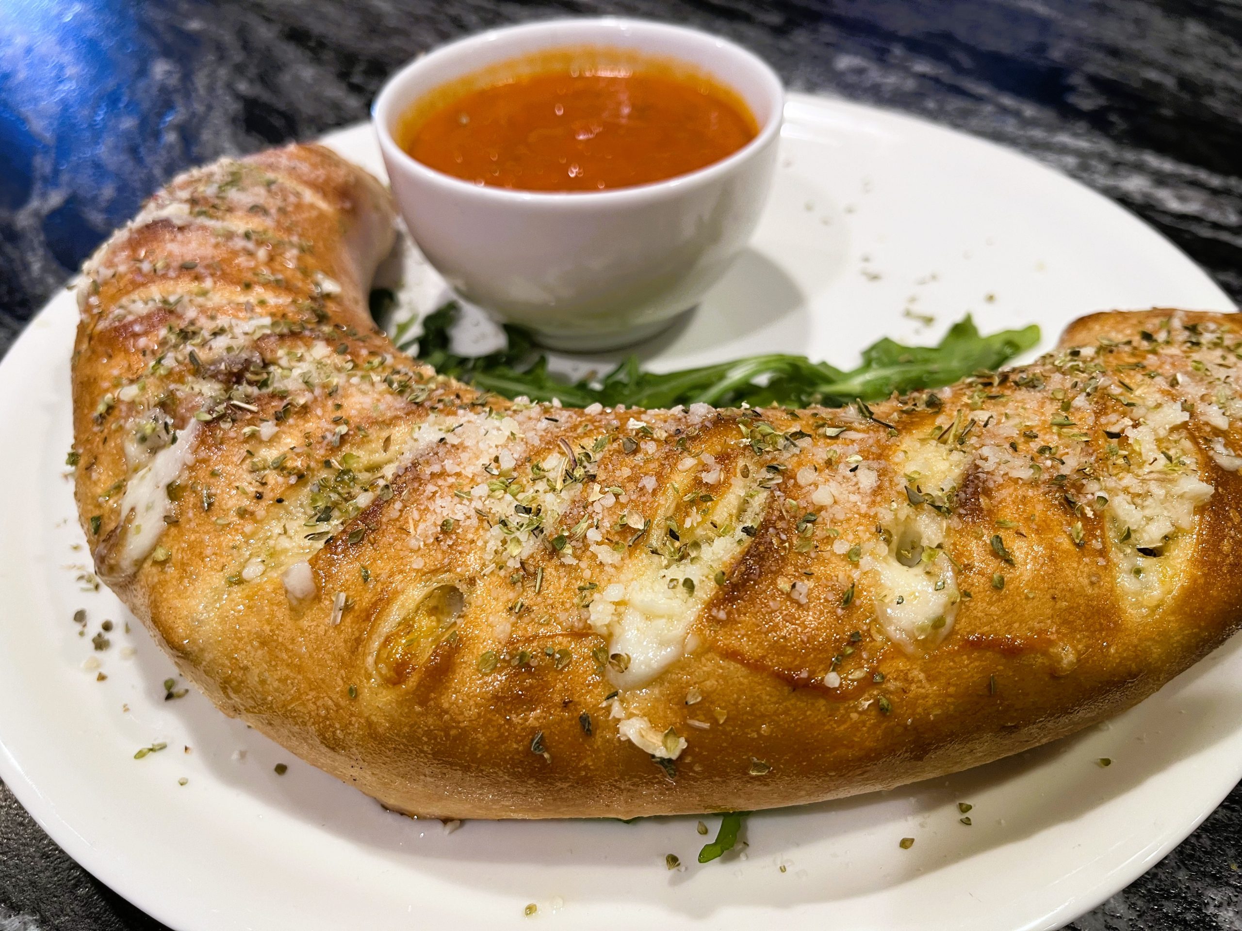 A crescent-shaped baked bread with herbs and cheese on a white plate, accompanied by a small bowl of tomato sauce on the side.