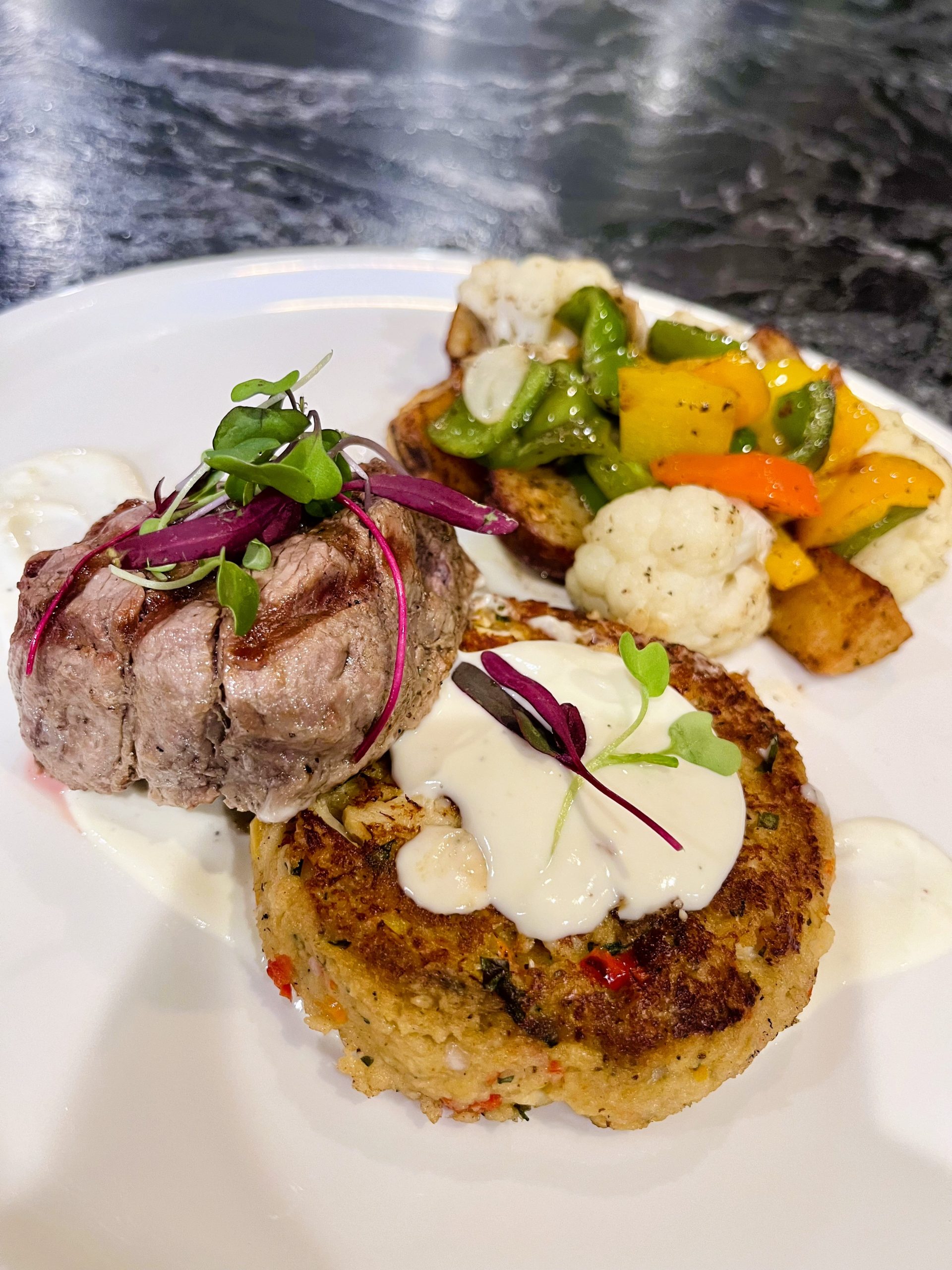 A plate with a steak medallion, garnished with microgreens, a crab cake with sauce, and a side of mixed vegetables, including cauliflower and bell peppers.