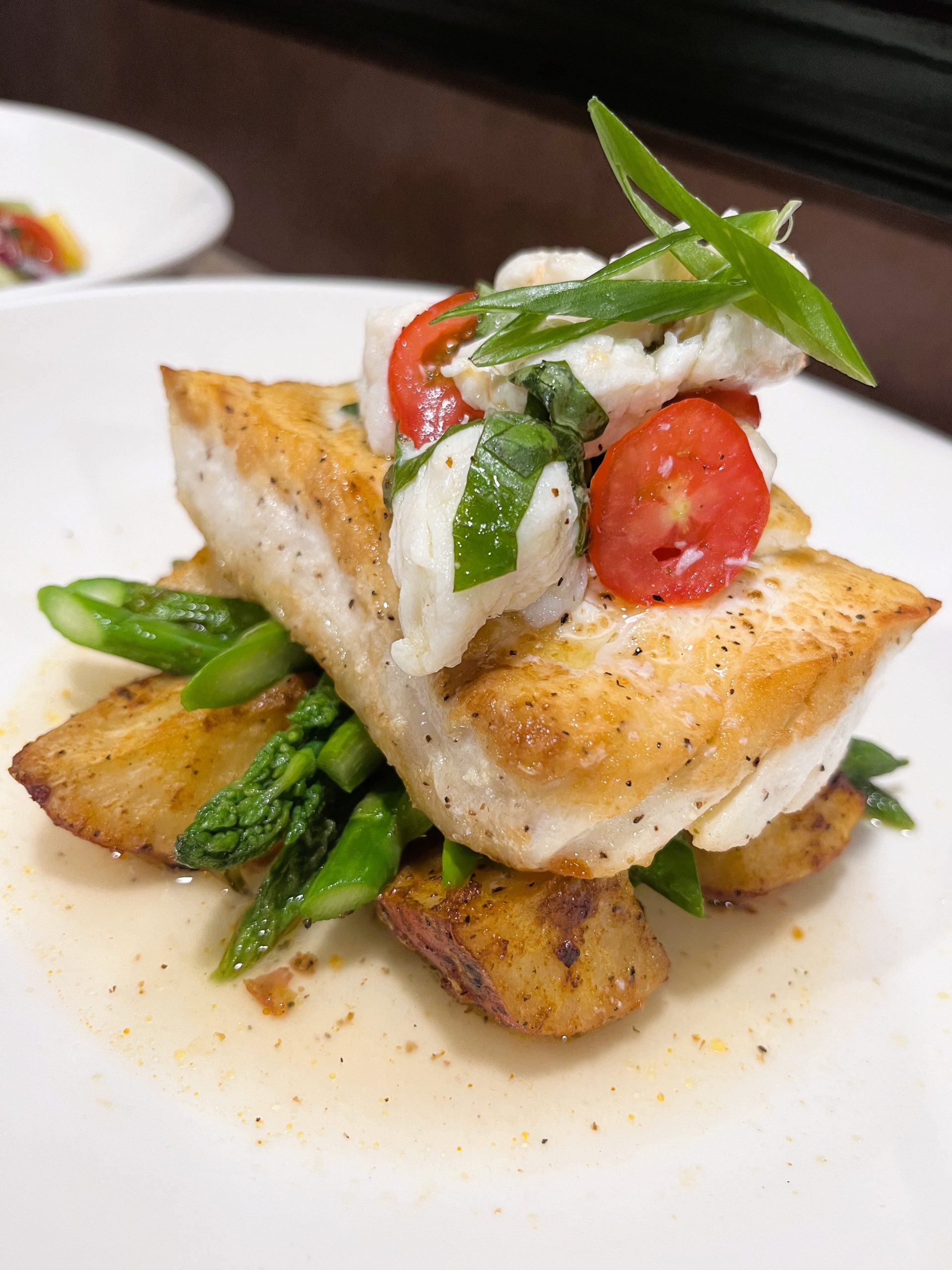 Grilled fish fillet on a bed of asparagus and roasted potatoes, topped with sliced red chili and green onions.