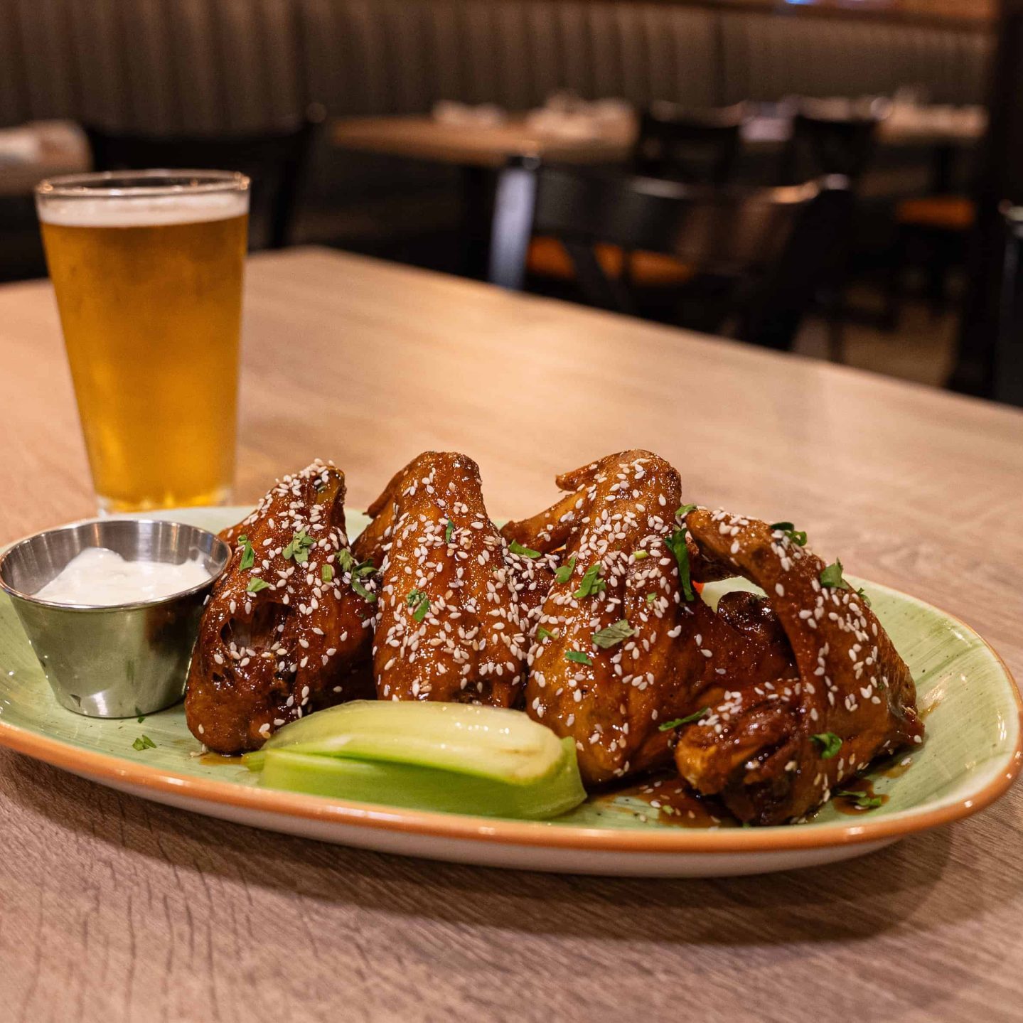 Plate of sesame-covered chicken wings with celery, ranch sauce, and a glass of beer on a wooden table in a restaurant. Empty chairs and tables are visible in the background.