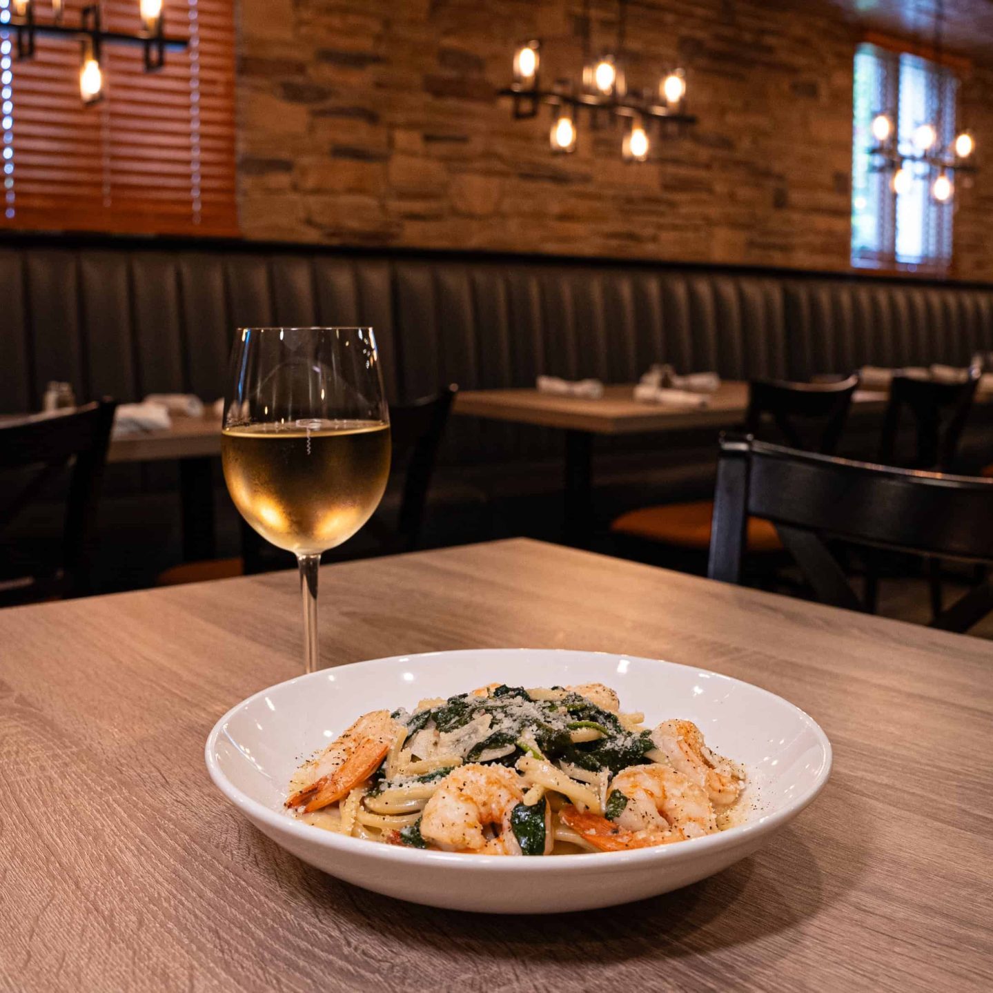 A plate of pasta with shrimp and vegetables served on a wooden table with a glass of white wine in a dimly lit restaurant.